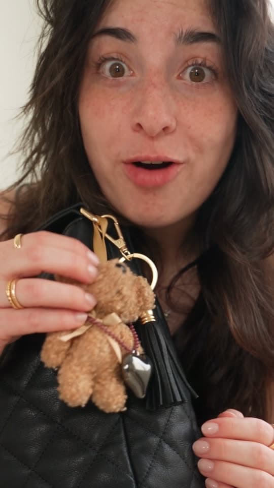 Cristina holding up a teddy bear charm with a silver heart and black tassel, clipped to a quilted black bag